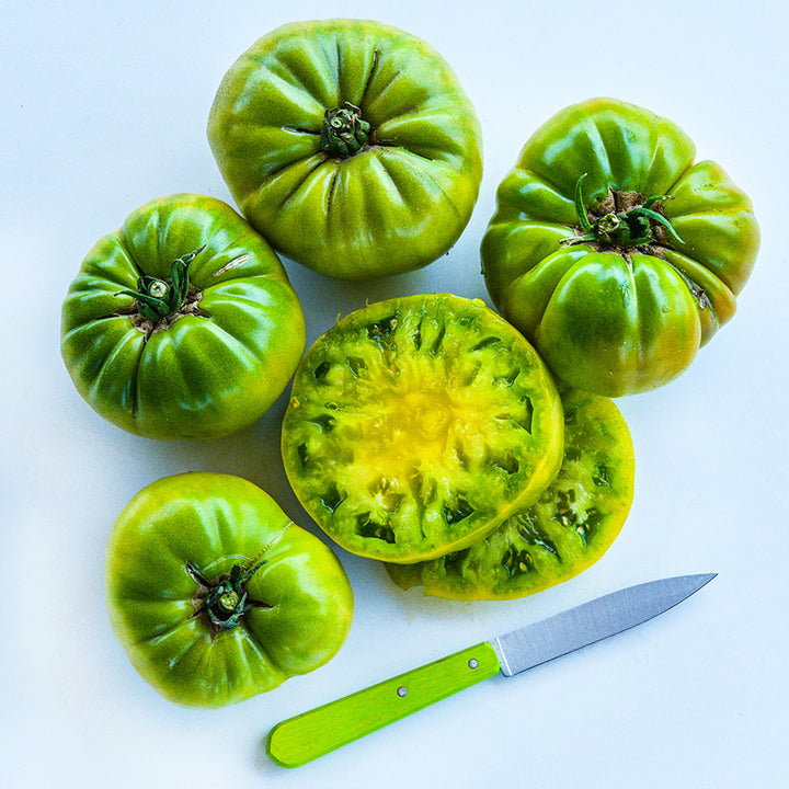 Overhead view of Green Giant Tomato slices surrounded by four tomatoes and a green handled knife.
