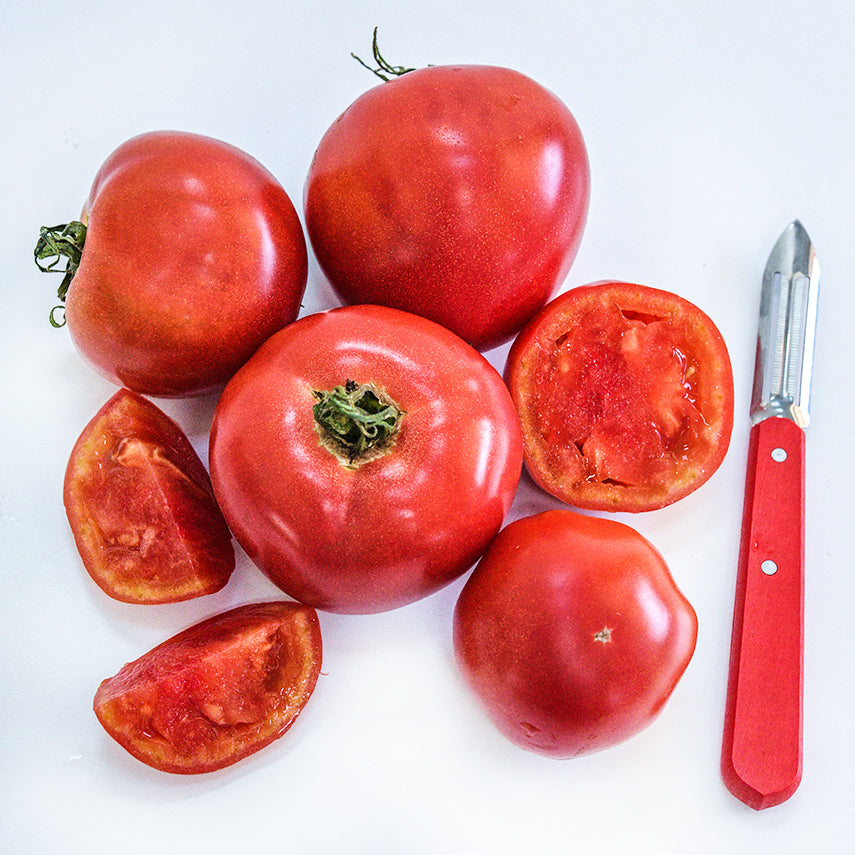 Whole, halved and quartered Ozark Pink Tomatoes with a red handled peeler on white background.