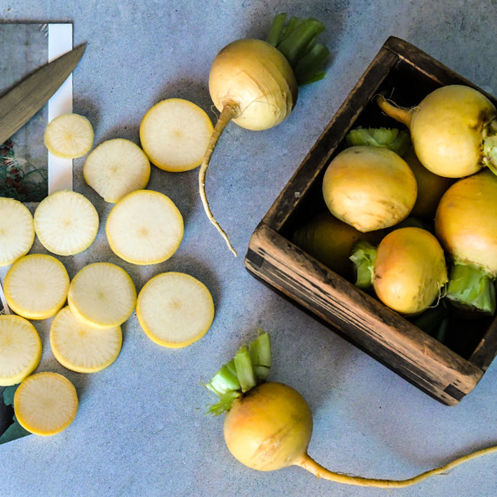 Whole and sliced yellow Aji Kogane Turnips with four in a Japanese wooden box on a light blue backdrop.