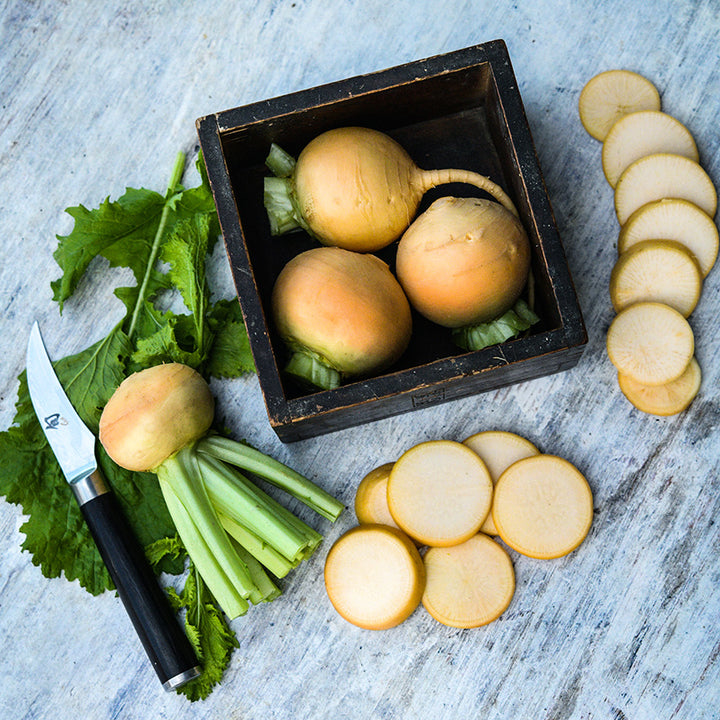 A trio of Aji Kogane Turnips in a wood box surrounded by sliced and whole turnips on a white wooden backdrop.