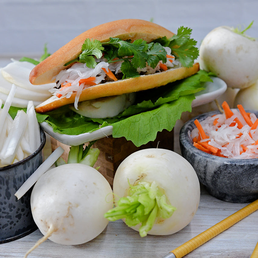 A Miyama Turnip Vegan Pho sandwich surrounded by cut, whole and shredded turnips on a light background.