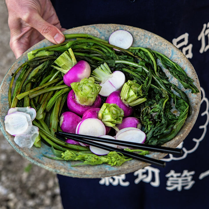 A man in a Japanese chef apron holding sauteed turnip greens and Nagasaki Akari Kabu Turnips.