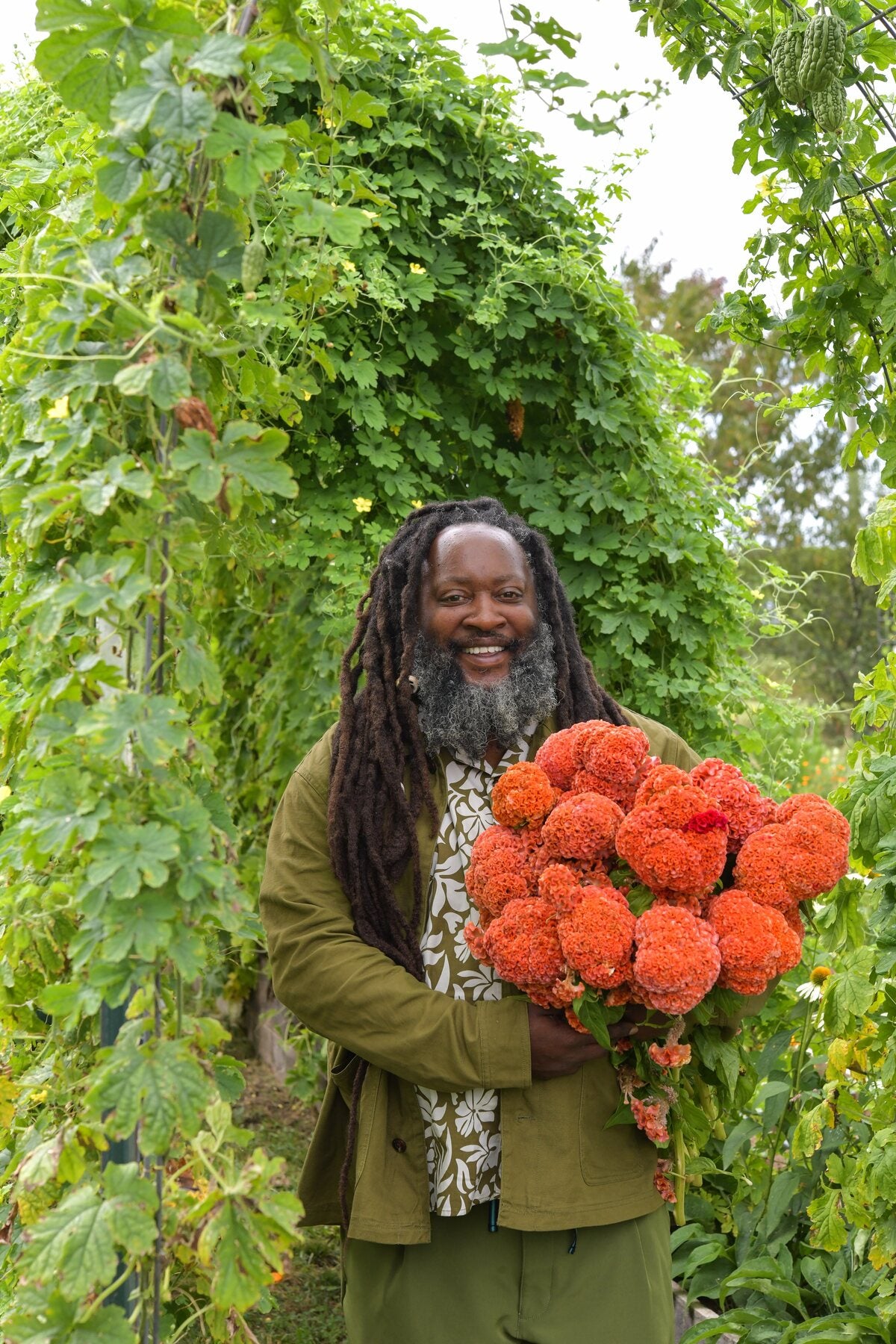 A smiling gardener holding an armful of orange celosia under an arch of vines