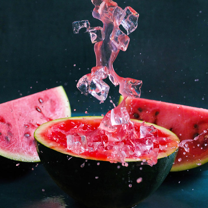Iced watermelon juice splashing on top of a cut Sugar Baby Watermelon on a dark blue background.