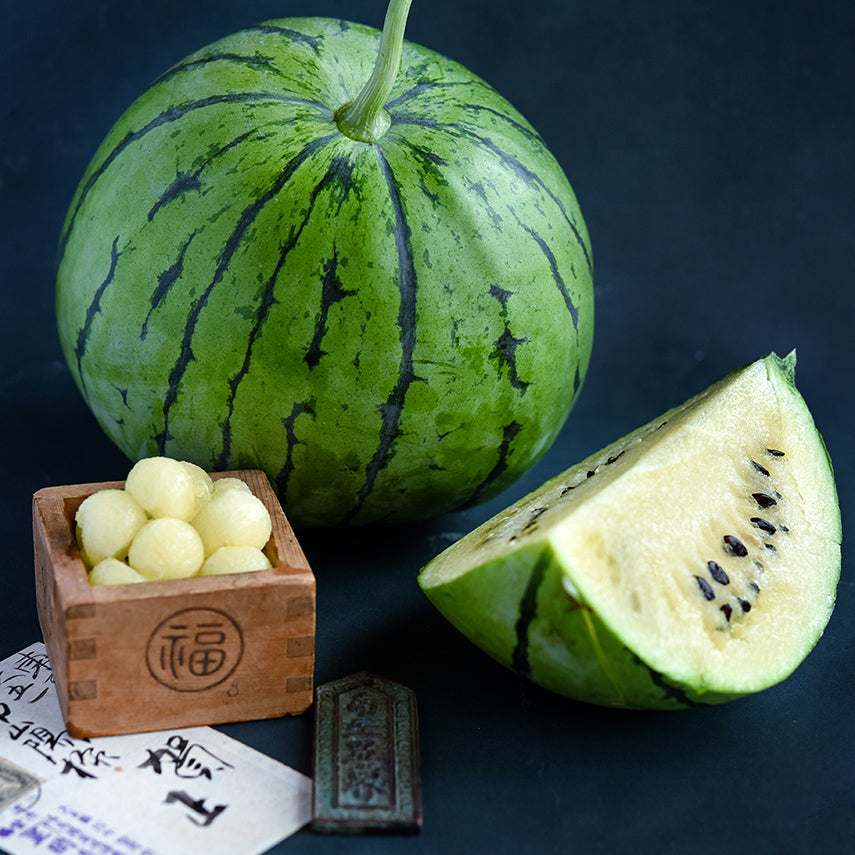 A whole Silver Yamato Watermelon with a quarter slice and melon balls in a wooden Japanese box on a jade background.