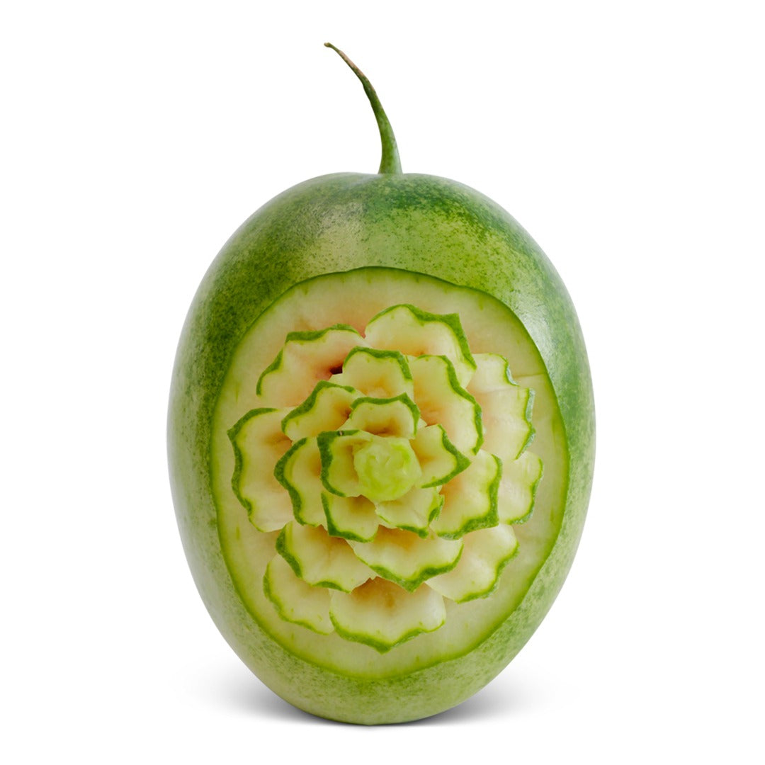 A small, oval Botswana Ice Cream watermelon with pale green skin, carved to reveal a layered floral pattern in the lighter flesh beneath.