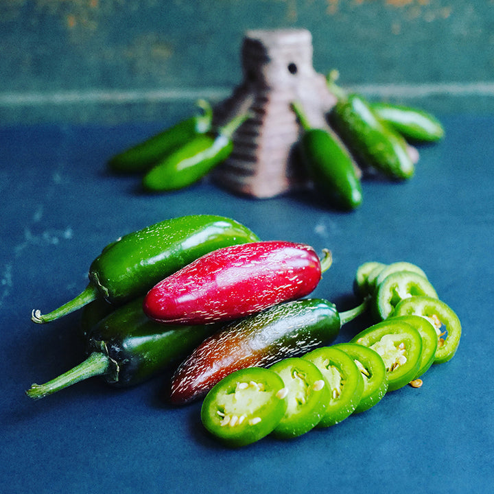 Zapotec Jalapeno, both red and green peppers and slices, on blue tabletop.