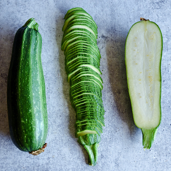 A trio of sliced and whole Nimba Zucchini Summer Squash on a speckled gray background.
