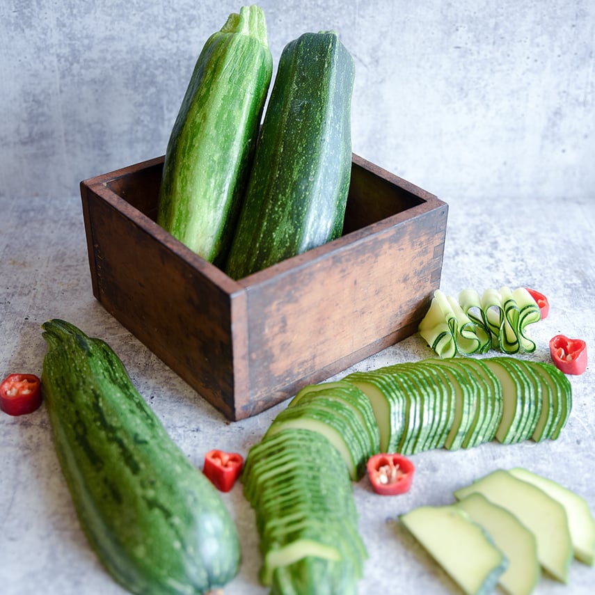 Thin sliced Nimba Zucchini Summer Squash with whole squash in a wooden box on a light background.