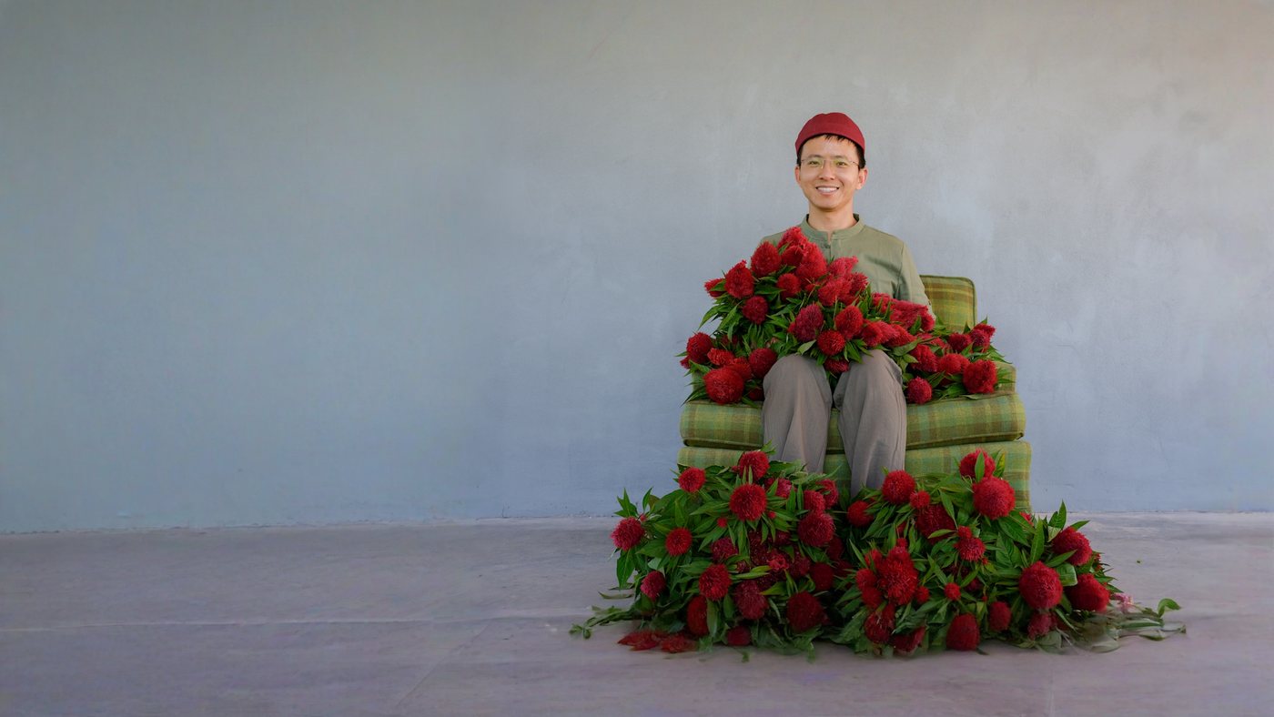 A smiling gardener in a red cap sitting in a green plaid armchair surrounded by red celosia flowers