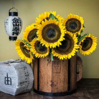An arrangement of Taiyo Sunflowers on a large wood bucket next to an antique book and lantern.
