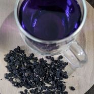 Black Goji Berry (Black Wolfberry) dried on a wooden plate with a cup of Goji berry tea in the background.