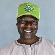 A happy smiling man wearing an Avocado Green Baker Creek Organic Cap against a grey wall.