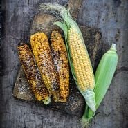 Grilled and seasoned Damaun KS Super Sweet Corn with raw cobs on a wooden background.