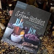 The Artisan Herbalist book by Bevin Cohen sits on a table surrounded by dried herbs and vintage bottles.