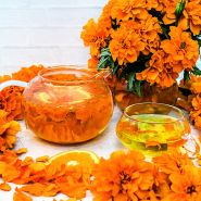 Safari Tangerine Marigolds with petals in a glass tea pot and cup surrounded by blooms on white.