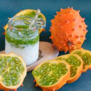 Sliced and whole Kiwano African horned jelly melons next to a yogurt pudding cup topped with melon on a dark blue background.
