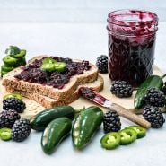 Apache Blackberry and jalapeno jam on toast surrounded by berry and peppers on a light background.