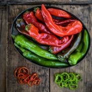 Red and green Txorixero Peppers in a bowl next to green and red slices on a wooden background.