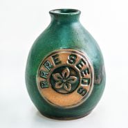 A single greenish blue glazed Handmade Stoneware Pottery Vase with a rareseeds logo on it on white.