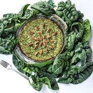 A spinach pie surrounded by fresh Bloomsdale Long Standing Spinach leaves and a fork.
