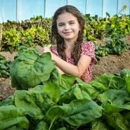 A smiling girl poses with large Gigante d' Inverno Spinach leaves in the greenhouse at Baker Creek.