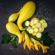 A trio of three Crookneck Early Golden Summer Squash with slices, blooms and leaves on a rust background.