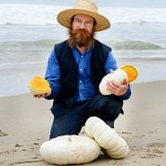 Man holding a cut Flat White Boer Pumpkin Winter Squash with a couple of pumpkins at his feet on the beach.