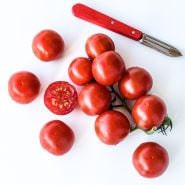 A cluster of cut and whole Chadwick Cherry or Camp Joy Tomatoes with a red handled peeler on white. 