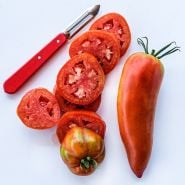 One whole and one sliced Jersey Devil tomato with a red handled peeler on a white background.