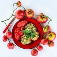 Baked Mortgage Lifter Tomatoes on bright red plate surrounded with tomatoes one a white background.