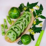 Green Zebra Tomatoes sliced on a wood serving dish surrounded by leaves and blooms.