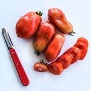 An arrangement of whole and sliced Martino's Roma Tomatoes with a red handled peeler on a white background.