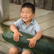 A laughing child holding a Giant Bullet Head Wax Melon seated on a porch.