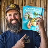 A smiling Baker Creek employee holding a couple of 2025 Whole Seed Catalogs.