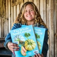 A smiling Baker Creek Employee holding out a couple of 2025 Whole Seed Catalogs.