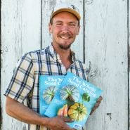 The Baker Creek farm manager smiles while holding a couple of 2025 Whole Seed Catalogs.