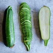 A trio of sliced and whole Nimba Zucchini Summer Squash on a speckled gray background.