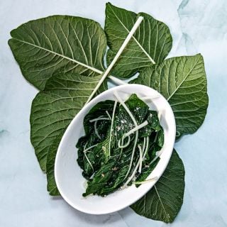 A bowl of cooked White Beauty Amaranth surrounded by fresh leaves on light green background.