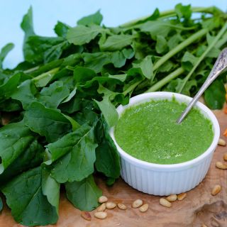 Fresh Common Arugula with arugula pesto surrounded with pinenuts on a wood cutting board.