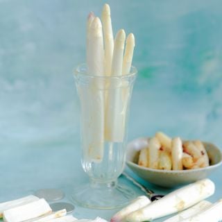 Precoce D'Argenteuil Asparagus spears in a glass with cooked and raw asparagus on light blue bakckground.
