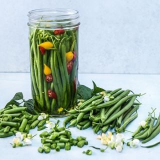 Pickled Opera Beans in a jar surrounded by whole and cut green beans, blooms and foliage.