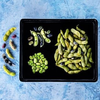 A black rectagular plate of steamed Tankuro Soybeans on a light blue background.