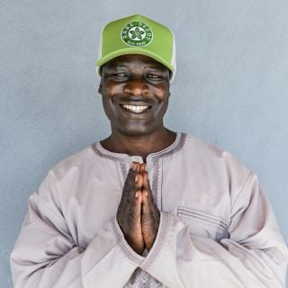 A smiling man holding his hands together while wearing an Avocado Green Baker Creek Organic Cap against a grey wall.