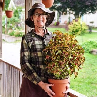 A smiling woman holding a potted Colocha Sunset Coleus plant on the front porch of the Seed Store.