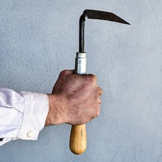 The Baker Creek blacksmith holding the hand-forged Cape Cod Right Handed Weeder against a plain gray wall.