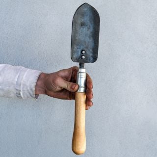 The Baker Creek blacksmith holding a Long Handled Trowel against a gray wall.
