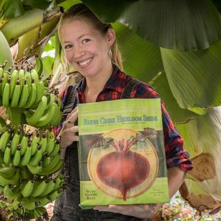 Deb smiles as she shows off the Baker Creek Handy Tote Bag by a banana tree.