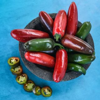 Nadapeno Peppers red, green, and brown shades in a bowl alongside fresh sliced  and whole peppers on a blue background.