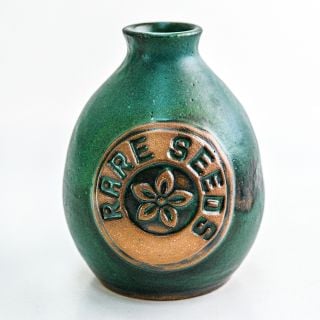 A single greenish blue glazed Handmade Stoneware Pottery Vase with a rareseeds logo on it on white.